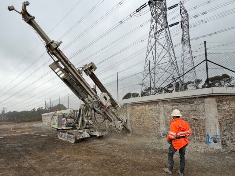 Track-mounted anchor drilling rig drilling into a shoring wall under transmission lines, with a KFM engineer inspecting, NSW